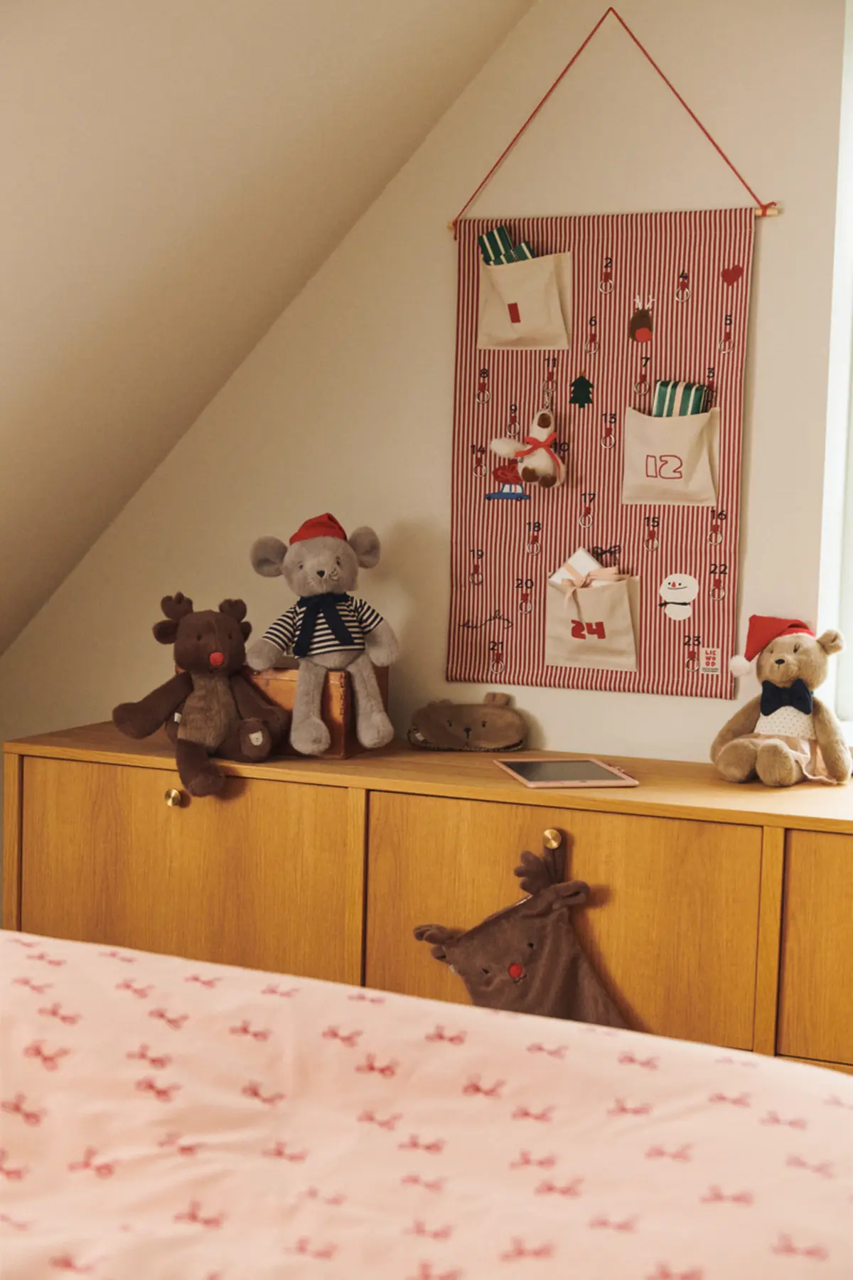 Blaise-Adventskalender an einer Holzwand über einer Kommode mit Kuscheltieren und einem Zora-Zeichenbrett.