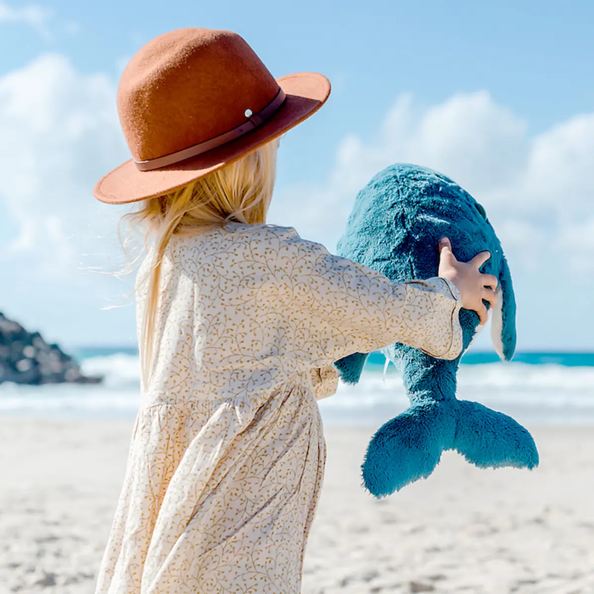 Kind trägt blauen Kuschelwal Hurley am Strand – O.B. Designs
